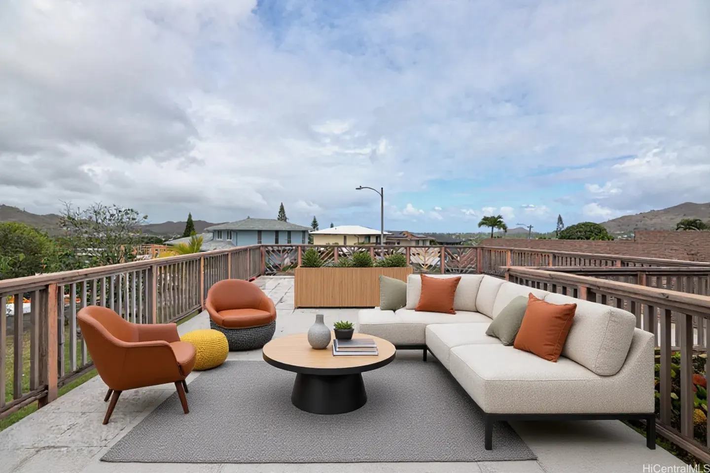 Outdoor rooftop patio with a white sectional, brown chairs, and a round coffee table on a gray rug. A wooden railing surrounds the patio.