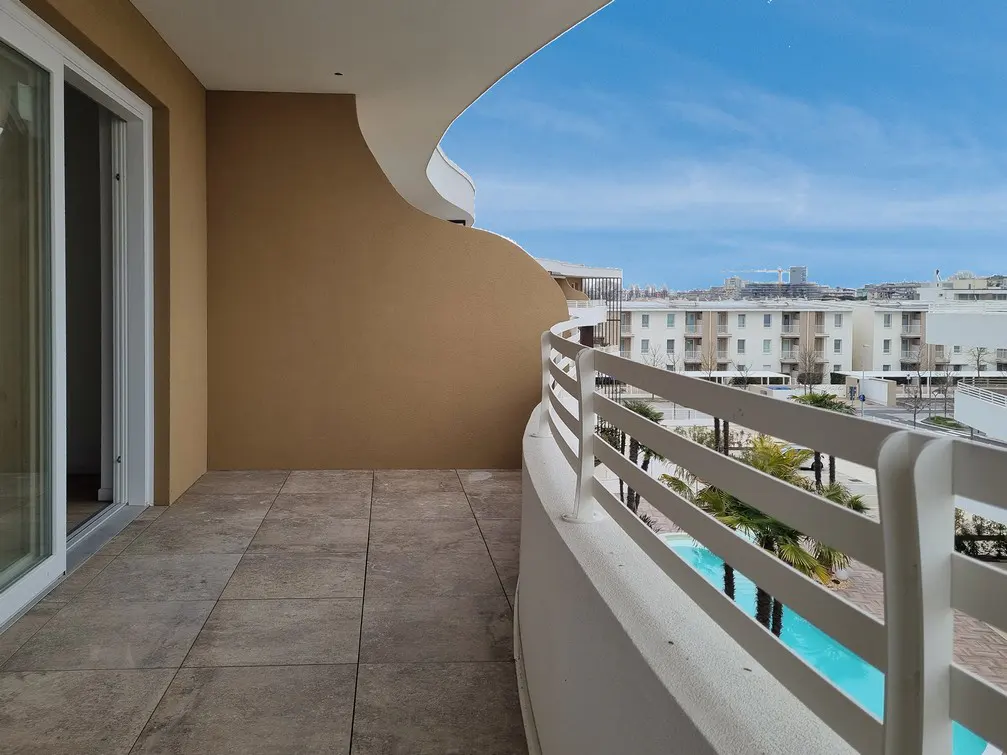 A curved balcony with white railings overlooks a city with a blue sky. The balcony has beige tile flooring and a sliding glass door.