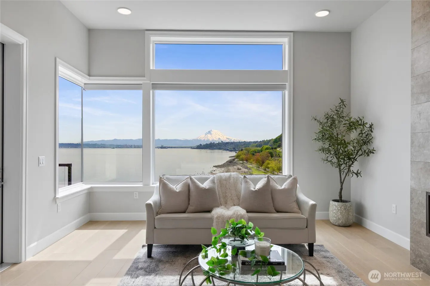Living room with a beige sofa, glass table, and a large window view of a lake and mountain.