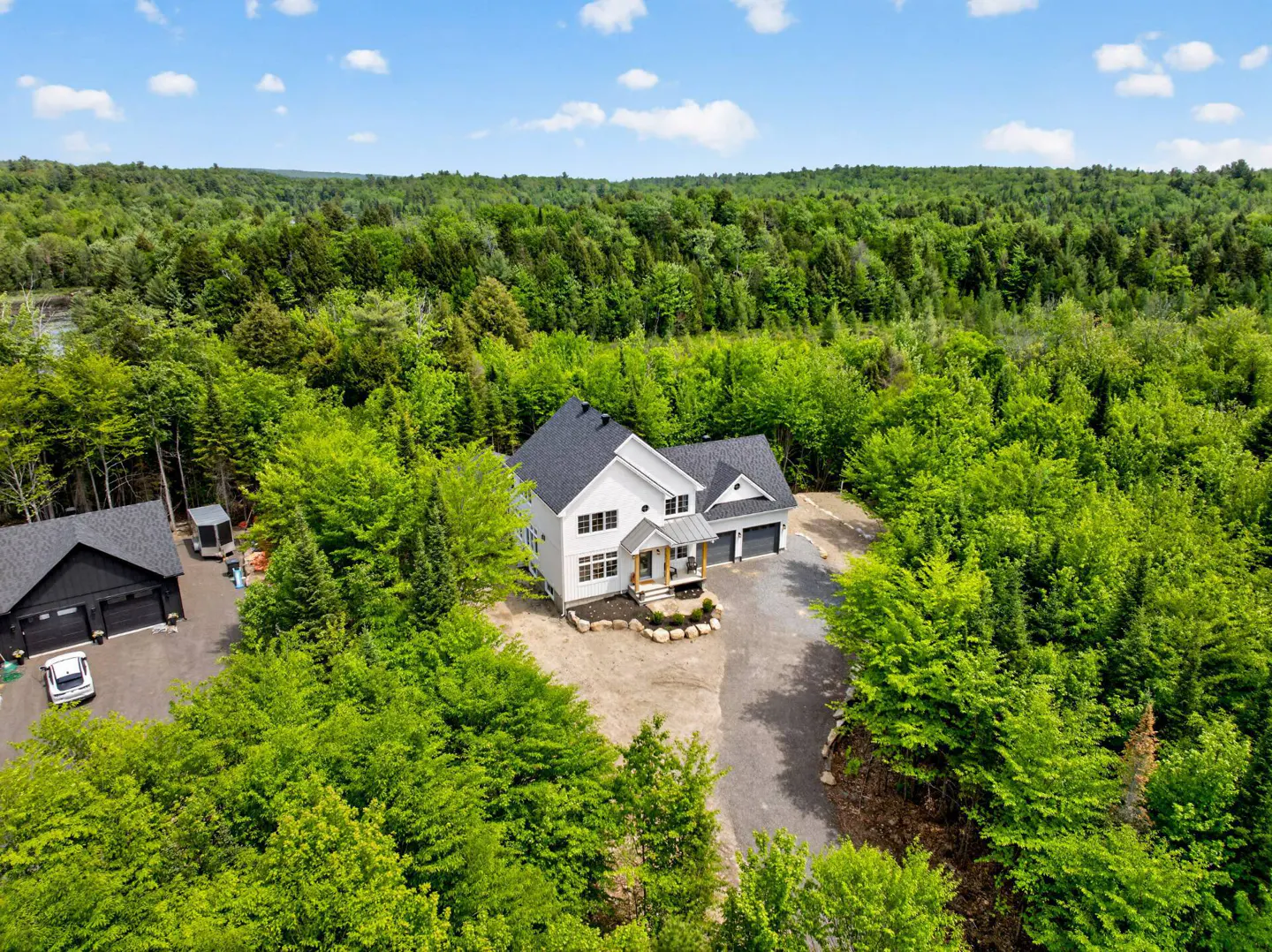 Aerial view of a white modern farmhouse with a dark roof, nestled in a lush green forest with a gravel driveway.
