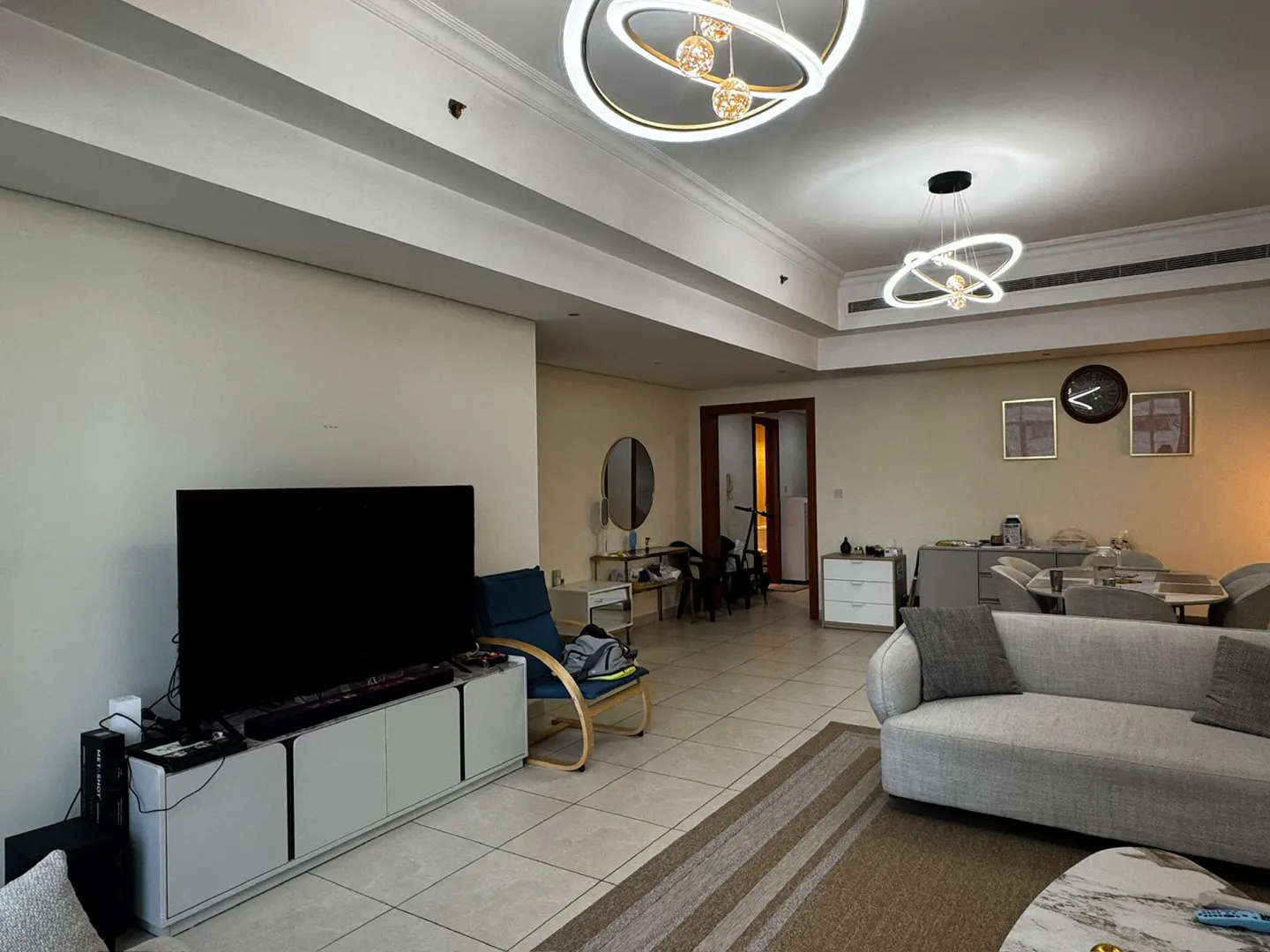 A living room with a TV, gray sofa, and modern lighting fixtures. A blue chair sits near a white media console.