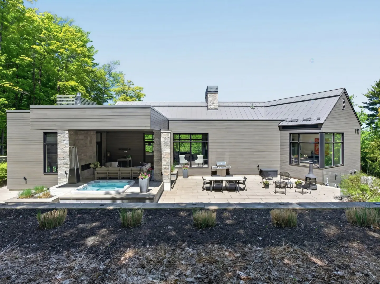Modern gray home with a stone-accented patio, outdoor hot tub, dining area, and lush forest backdrop.