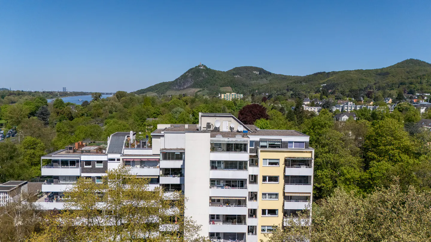 View of a multi-story apartment building with balconies, surrounded by green trees and hills under a clear blue sky.