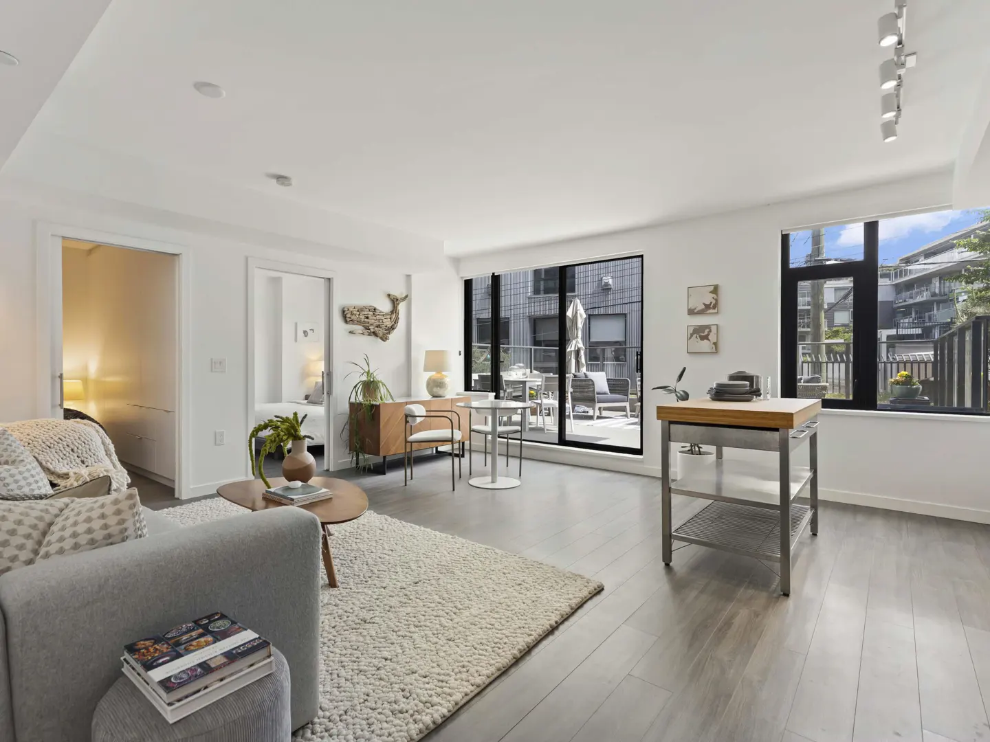 Bright, modern living room with gray sofa, wood floors, and white walls. A sliding glass door leads to a patio with outdoor seating.