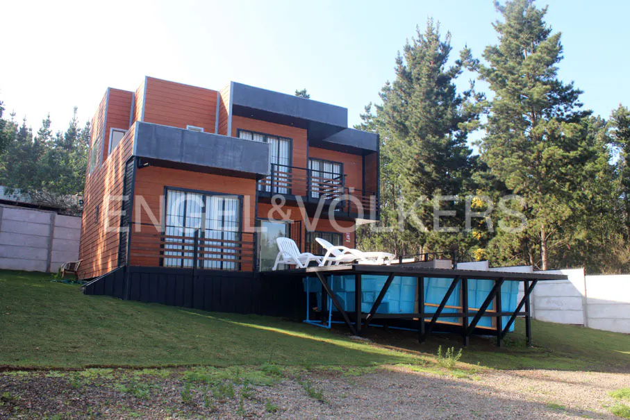 Modern two-story house with a pool, wood siding, and black trim. Two white lounge chairs sit on the deck. Trees in the background.