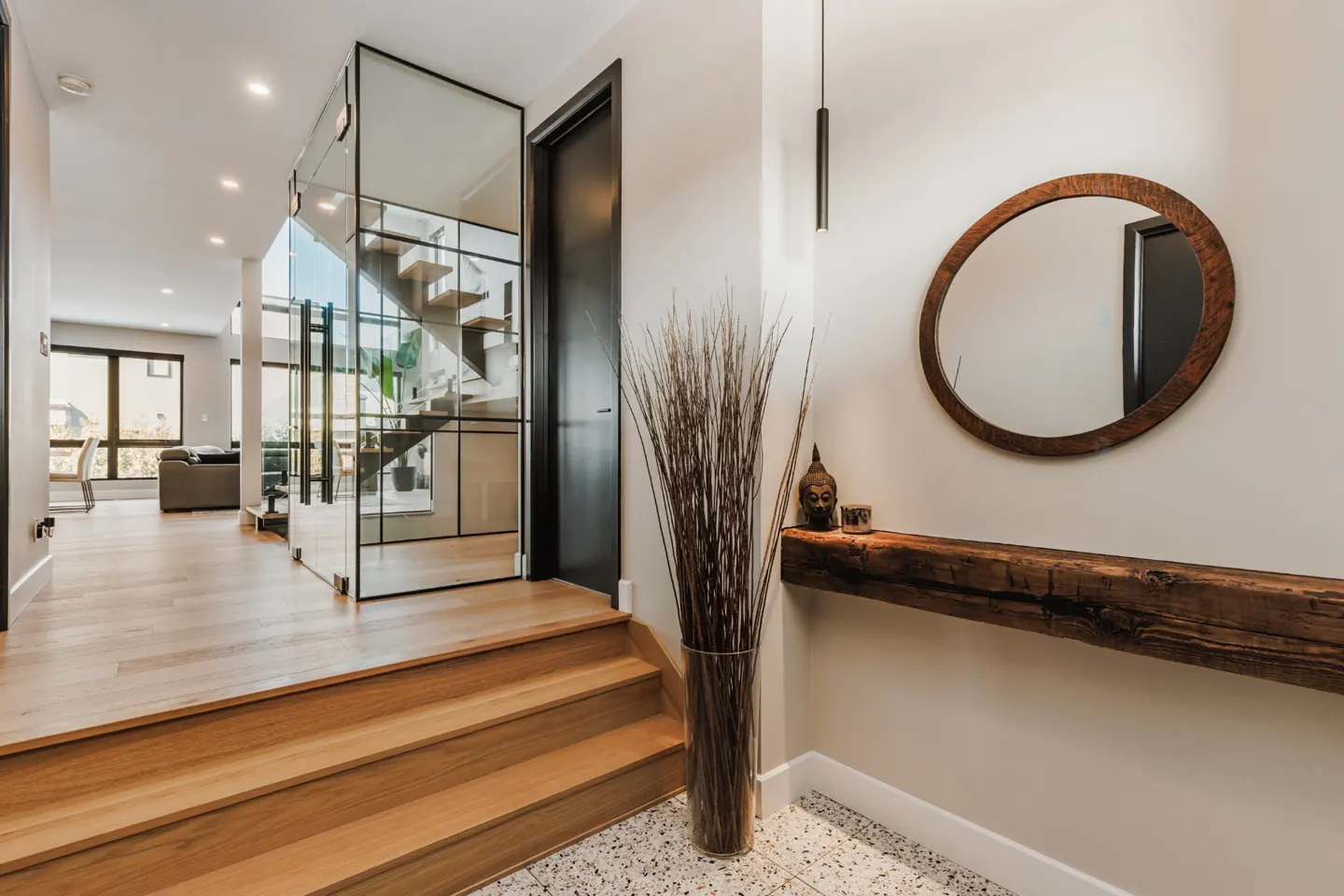 Modern home foyer with wood floors, glass-enclosed staircase, and a rustic wood shelf with a round mirror above.