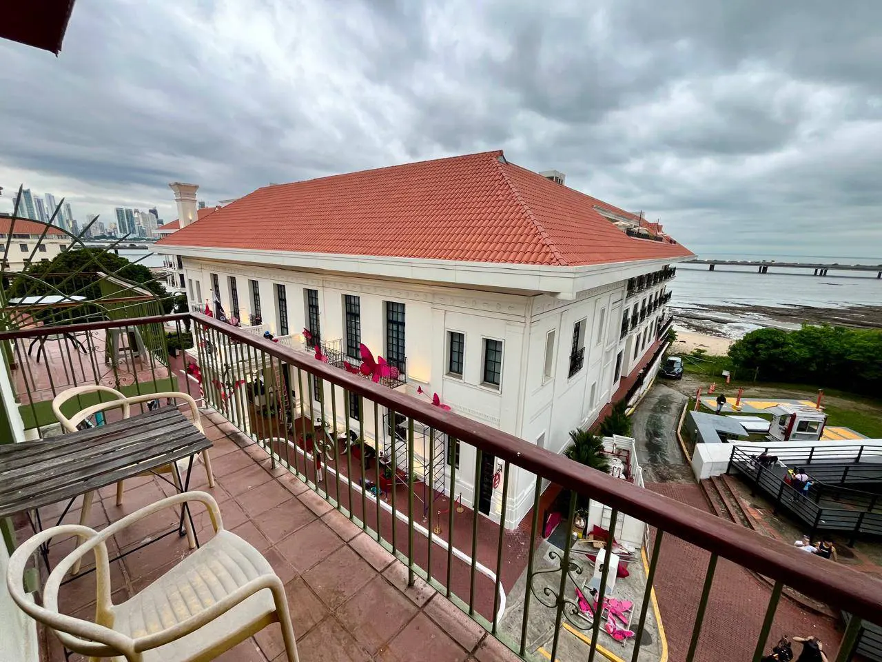 View from a balcony overlooking a large white building with a red tile roof, coastal scenery, and a cloudy sky.