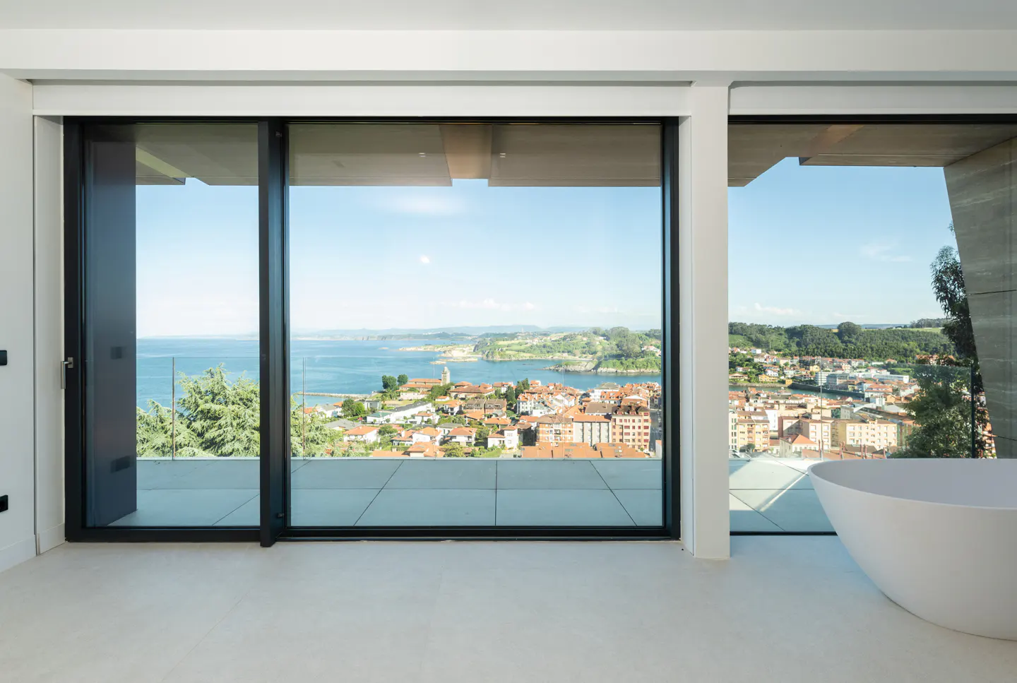 Modern bathroom with a white freestanding tub, floor-to-ceiling windows, and a panoramic view of a coastal town and sea.