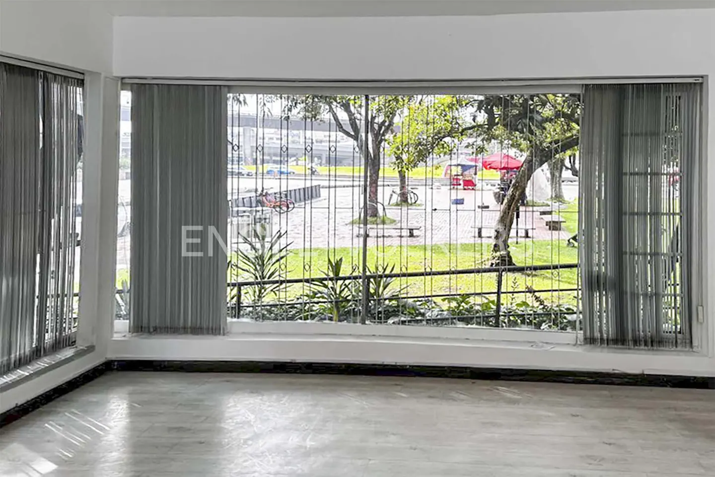 Empty room with a large window overlooking a park with trees, benches, and a red food stand. Gray blinds partially cover the window.