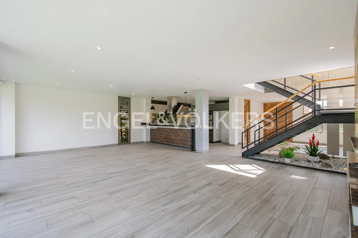 Open-plan living area with light wood-look tile floors, a modern kitchen, and a black metal staircase over a stone garden.