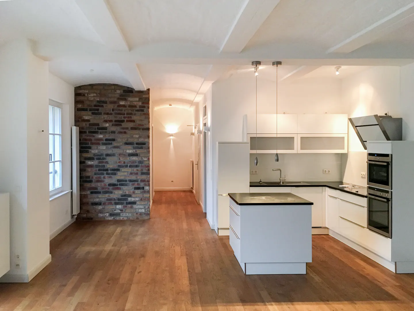 Bright, open apartment with hardwood floors, white walls, and a modern white kitchen with a black countertop island. A brick accent wall adds texture.