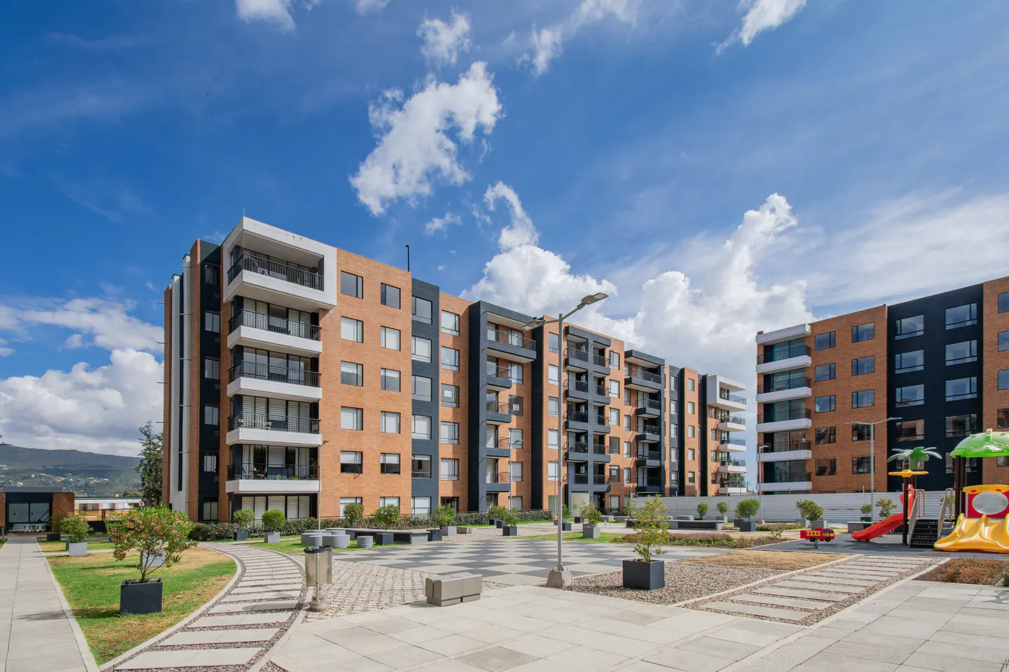 Modern apartment buildings with brick and dark gray accents, a playground, and landscaped grounds under a blue sky.