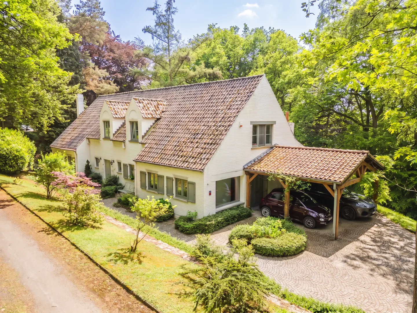 Two-story white house with a brown tile roof and a two-car carport, surrounded by green trees.