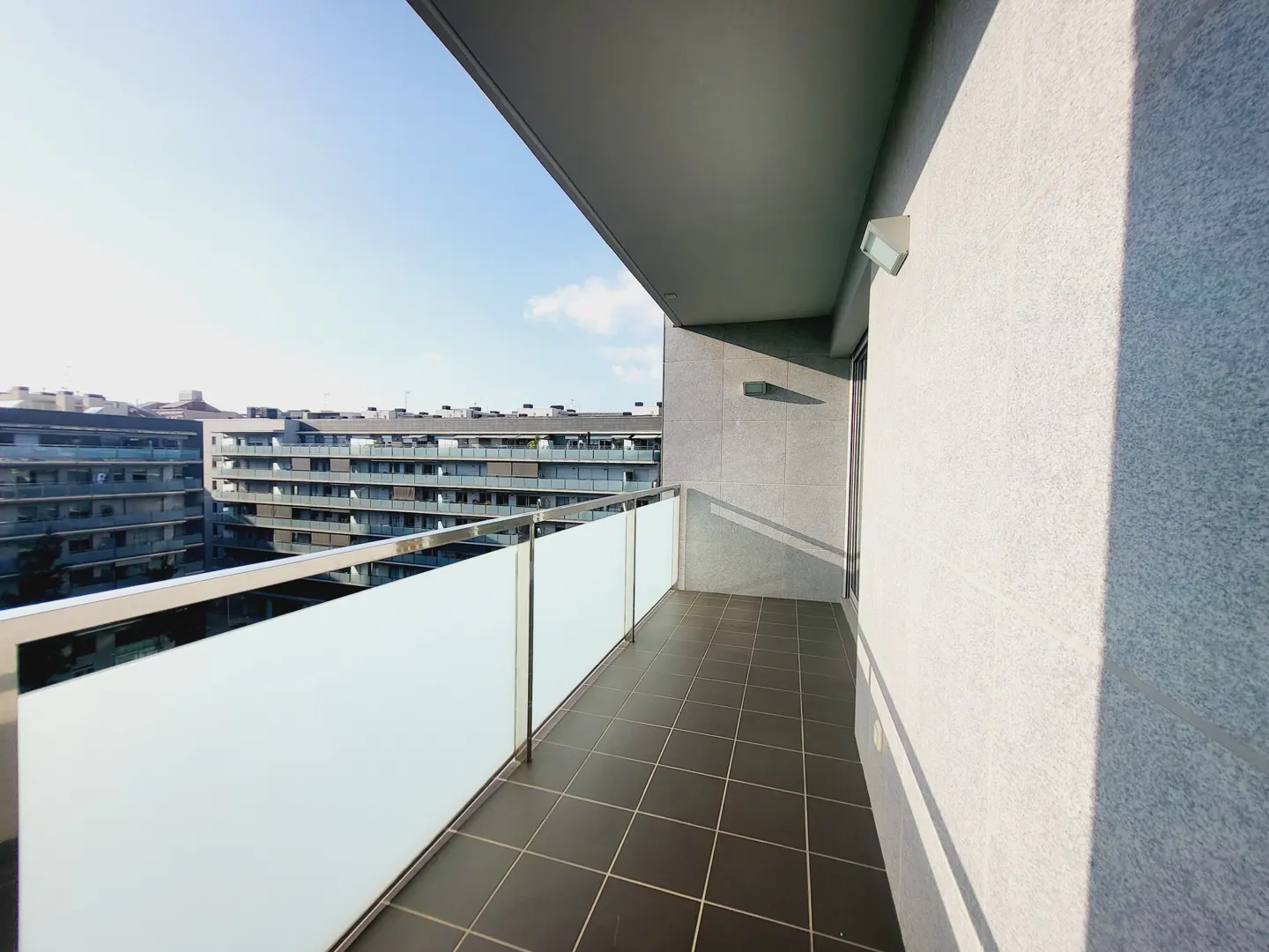 A modern balcony with dark tile flooring, a frosted glass railing, and a view of nearby apartment buildings.