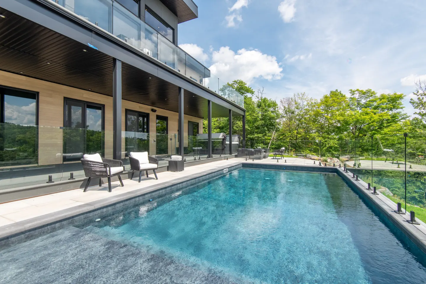 Modern home exterior with a blue swimming pool, glass railings, patio furniture, and lush green trees in the background.