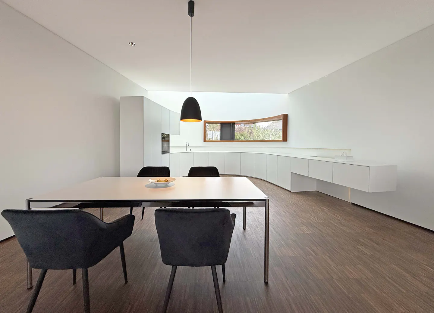 Modern dining area with white walls, wood floors, and black chairs around a white table. A black pendant light hangs above. A curved window is in the background.