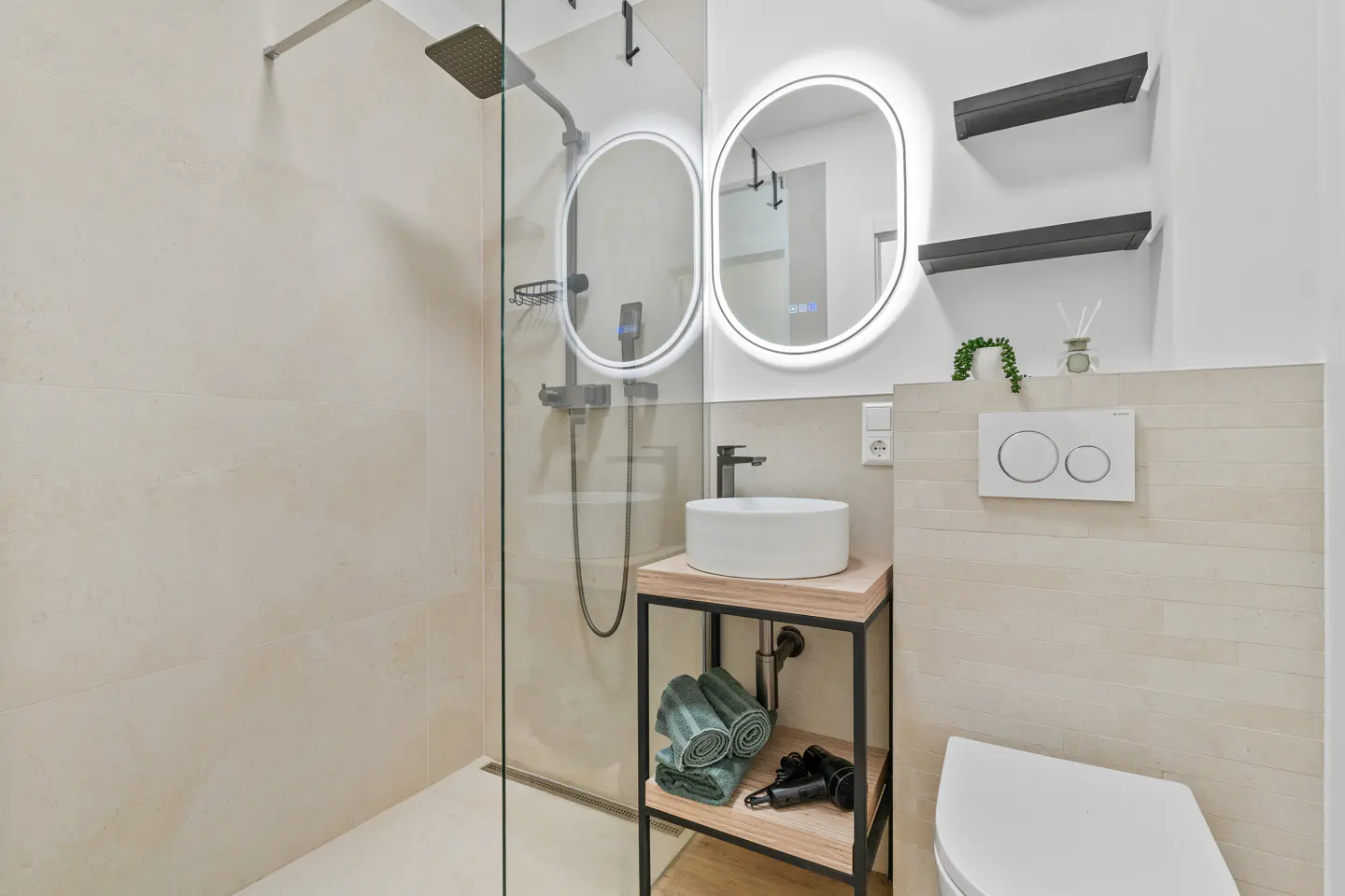 Modern bathroom with a glass shower, white sink on a wooden stand, and oval mirrors with LED lighting. Toilet and dark shelves are visible.