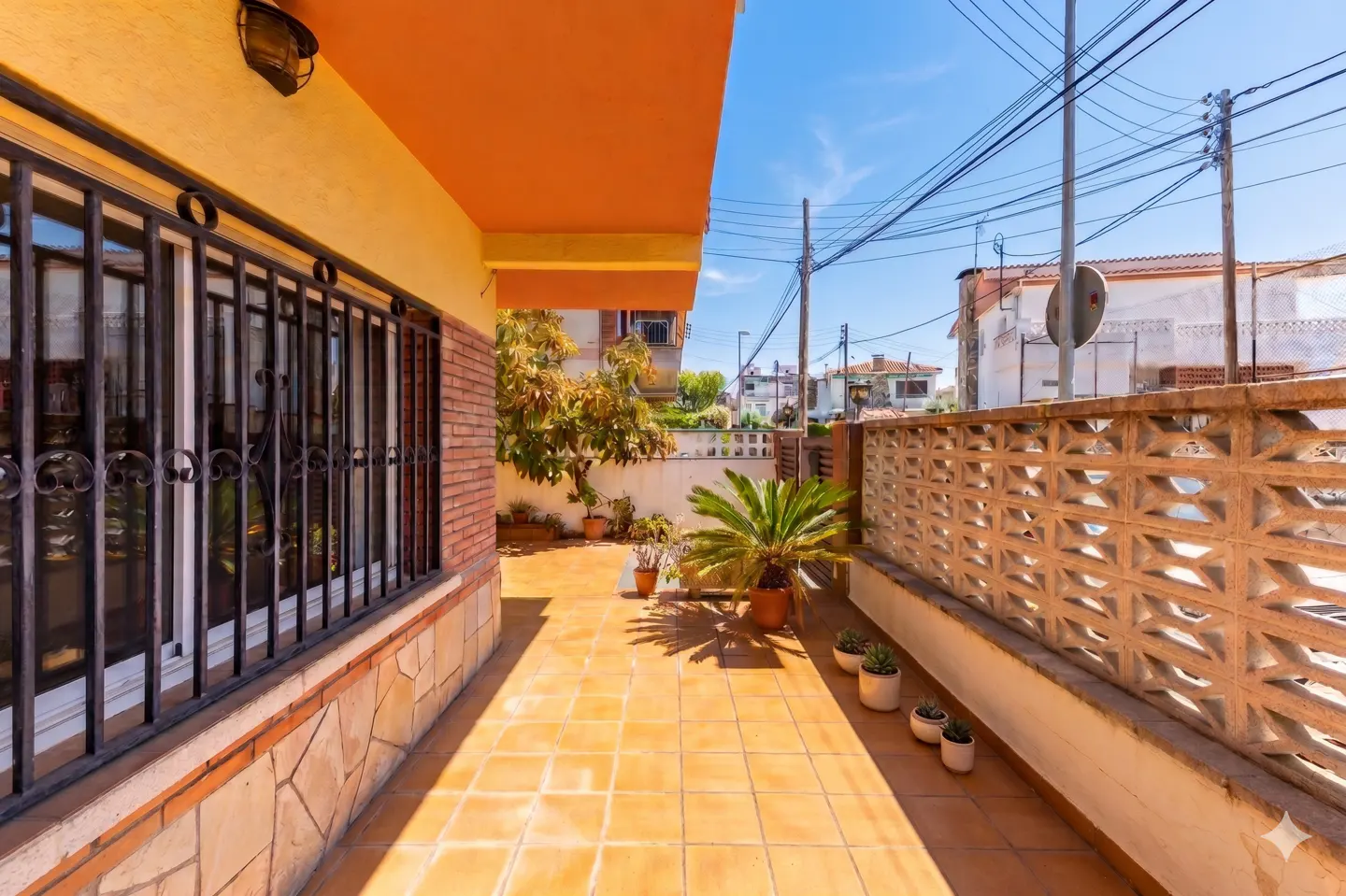 Outdoor patio with orange tile flooring, a brick wall, and a window with black bars. Potted plants line the patio.