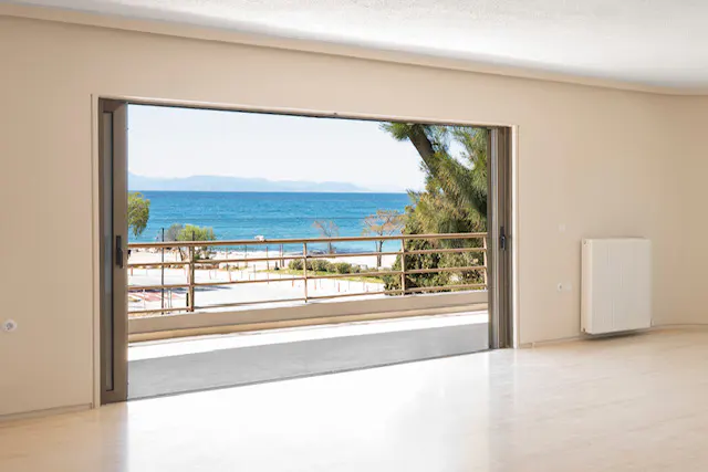 A bright room with an open sliding glass door reveals a balcony overlooking a beach and ocean view.