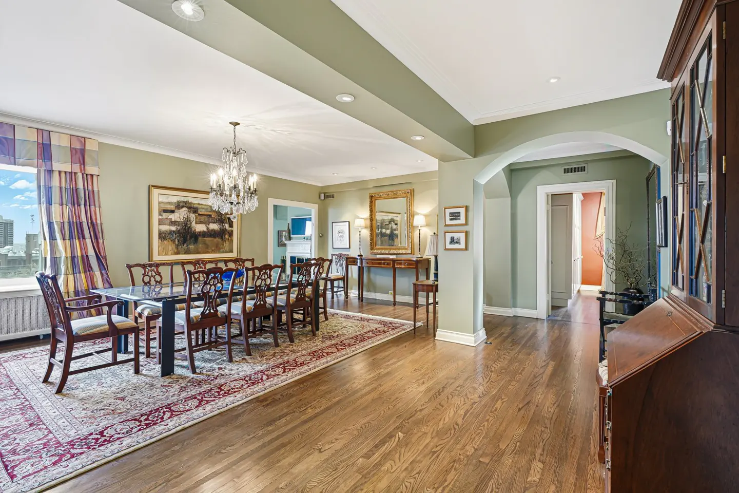 A dining room with hardwood floors, a long table with chairs, and a crystal chandelier. The walls are light green, and there is a large window with patterned curtains.
