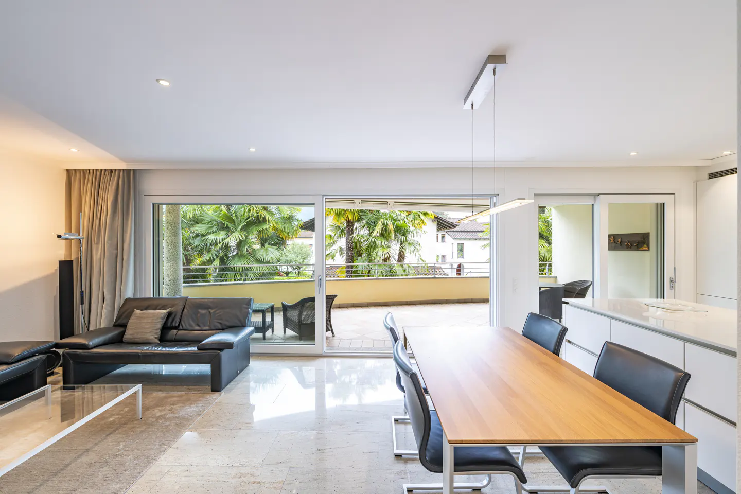 Bright, open-concept living space with a black leather sofa, wood dining table, and a balcony with palm trees.