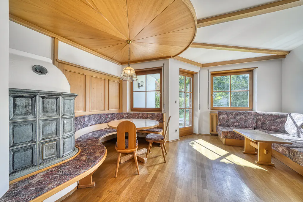 Dining area with a circular wood ceiling, tiled stove, floral built-in benches, wooden tables, and hardwood floors.