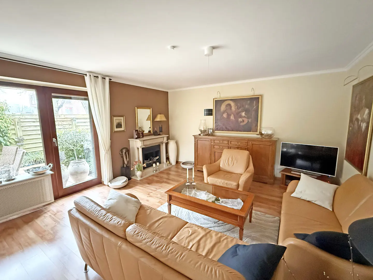 A living room with tan leather sofas, a fireplace, and a wooden cabinet with a painting above it. A glass door leads to a garden.