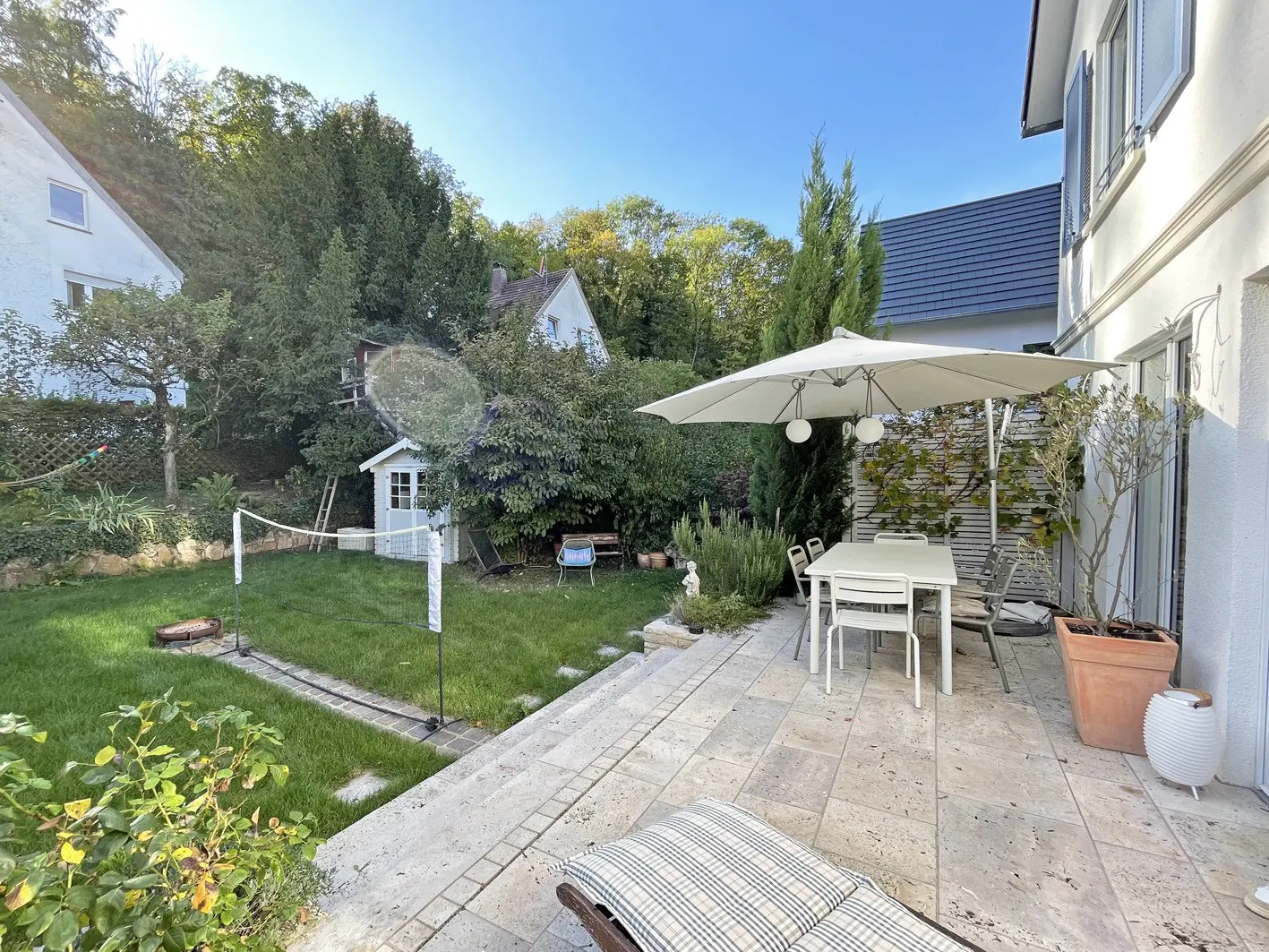 Backyard patio with a white table and umbrella, a green lawn with a volleyball net, and a small white playhouse.