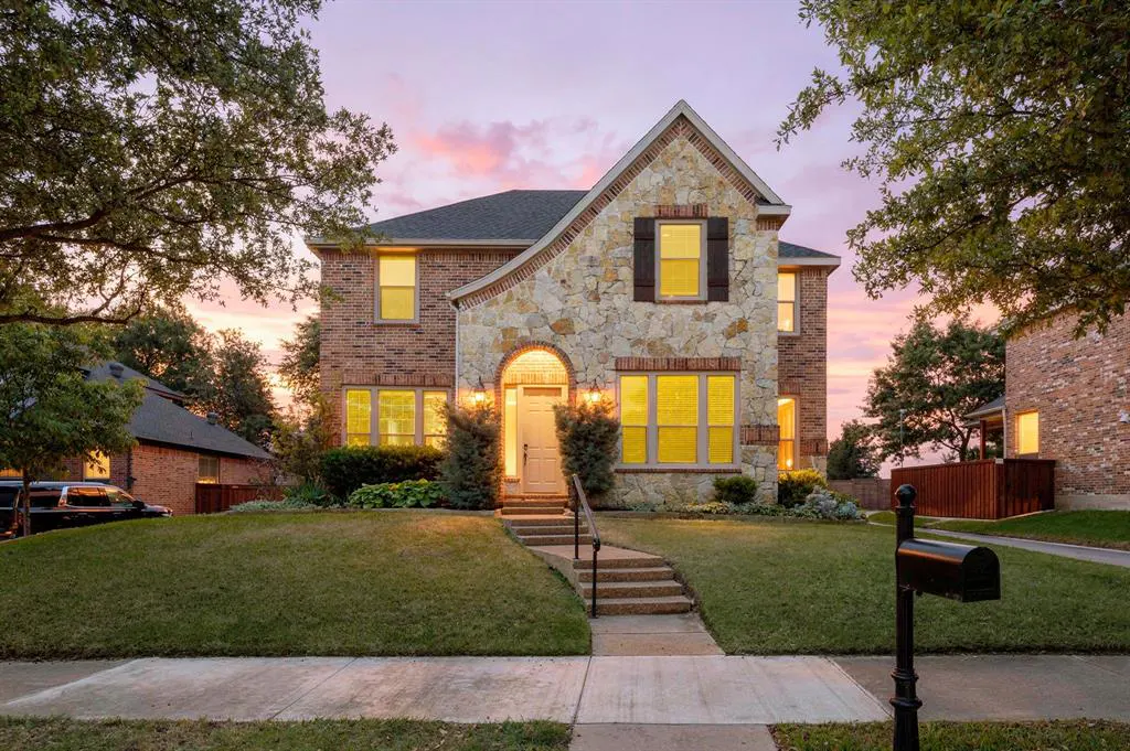 Two-story house with brick and stone facade, illuminated windows, and a manicured lawn at dusk.