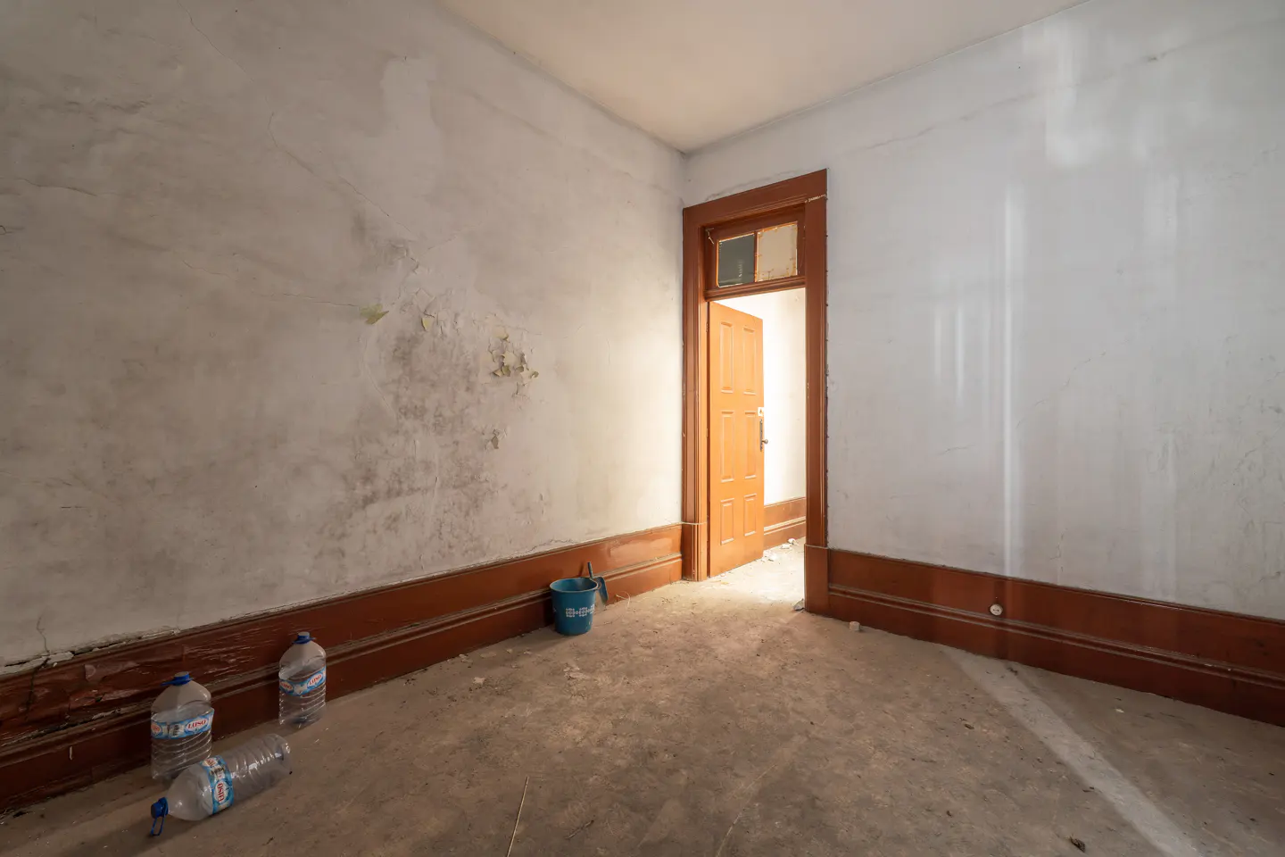 Empty room with peeling white walls, brown trim, and an open wood door. Water bottles and a blue bucket sit on the floor.