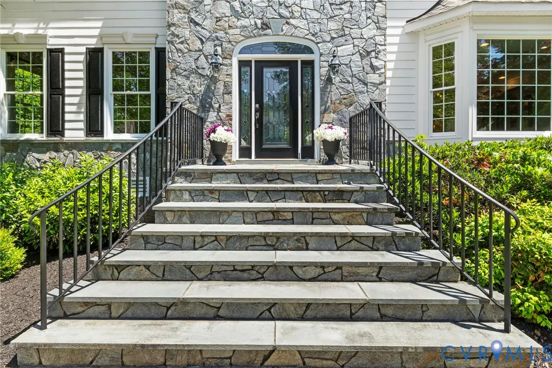 Stone steps lead to a black front door framed by stone. Black railings, flower pots, and windows flank the entrance.