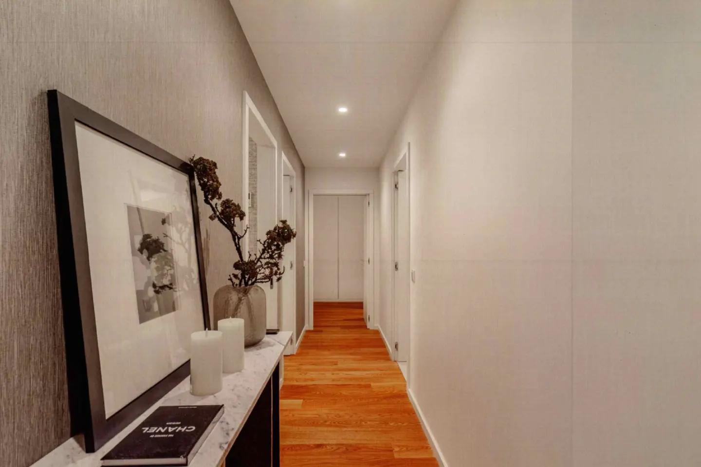 Hallway with wood floors, white walls, and recessed lighting. A console table displays a framed picture, vase, candles, and a book.