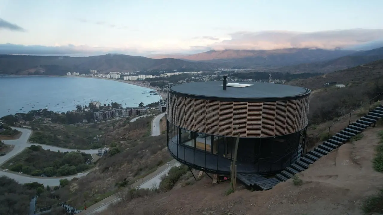 Modern round house with wood slats on a hill overlooking a bay and town. Stairs lead up the hill to the house.