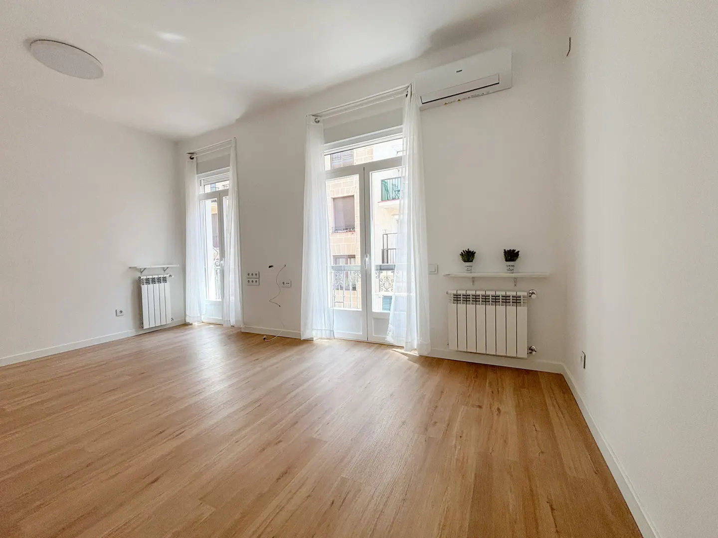 Bright, empty room with wood floors, white walls, and two windows with white curtains. A radiator sits beneath each window.