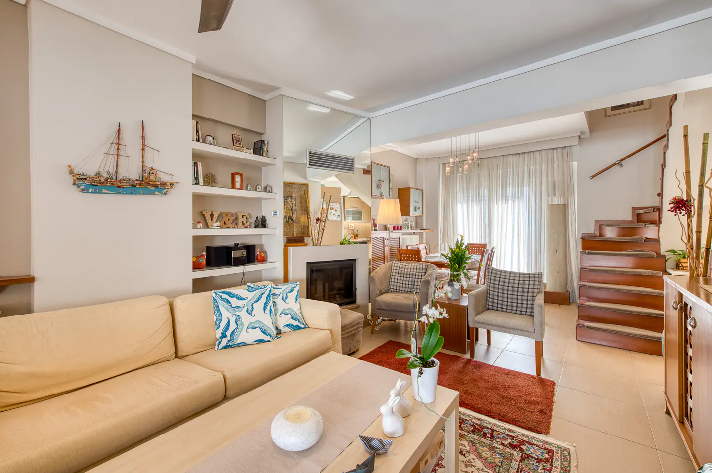 A bright living room with a beige sofa, blue pillows, a fireplace, and a wooden staircase in the background.