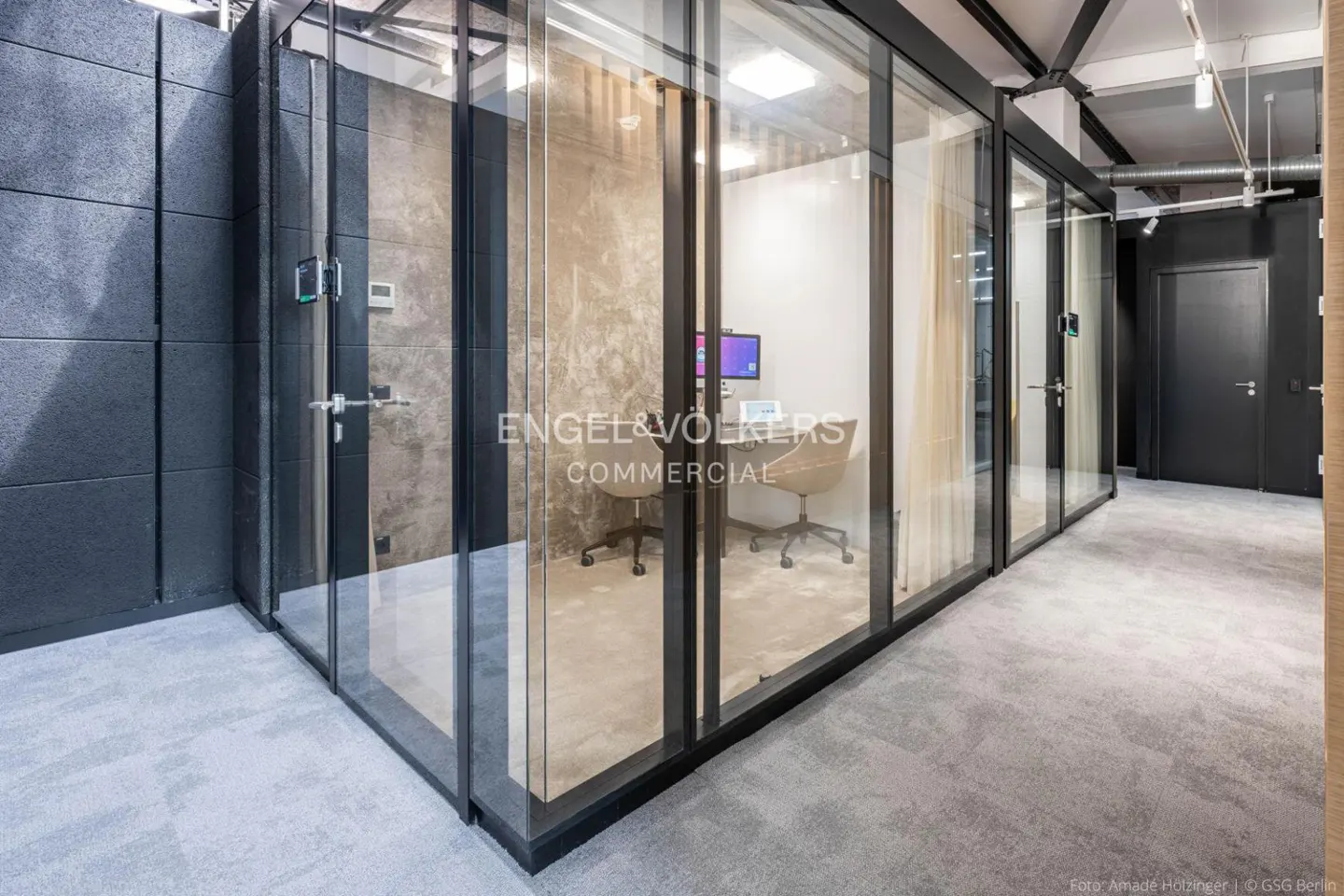 Office interior with glass-walled rooms, gray carpet, and black accents. Desks and chairs are visible inside the rooms.