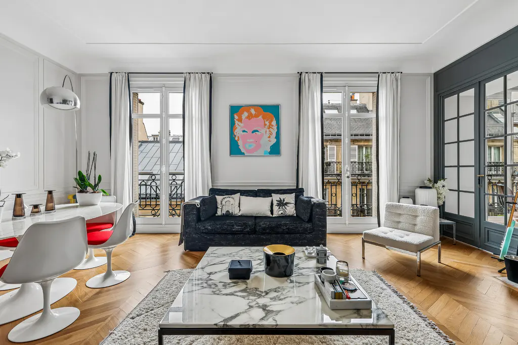 Bright living room with herringbone wood floors, white walls, and large windows overlooking Parisian buildings. A marble coffee table sits on a light rug.