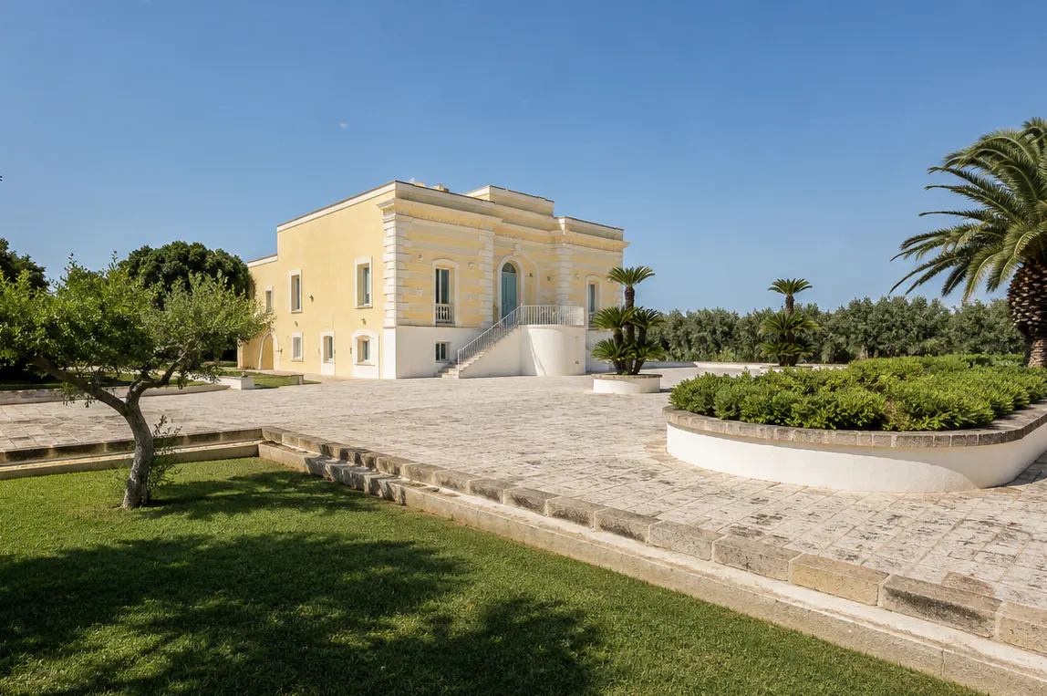 A pale yellow Mediterranean villa with a stone staircase, set in a sunny courtyard with palm trees and green lawns.
