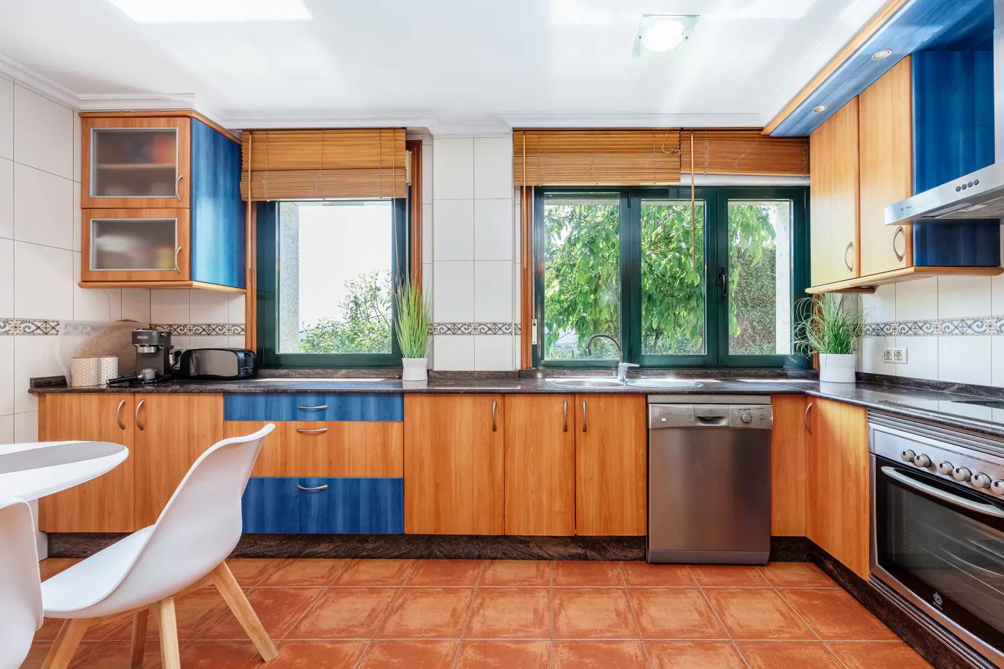 Bright kitchen with wood and blue cabinets, stainless steel appliances, and terracotta tile floor. Two windows with bamboo blinds overlook greenery.