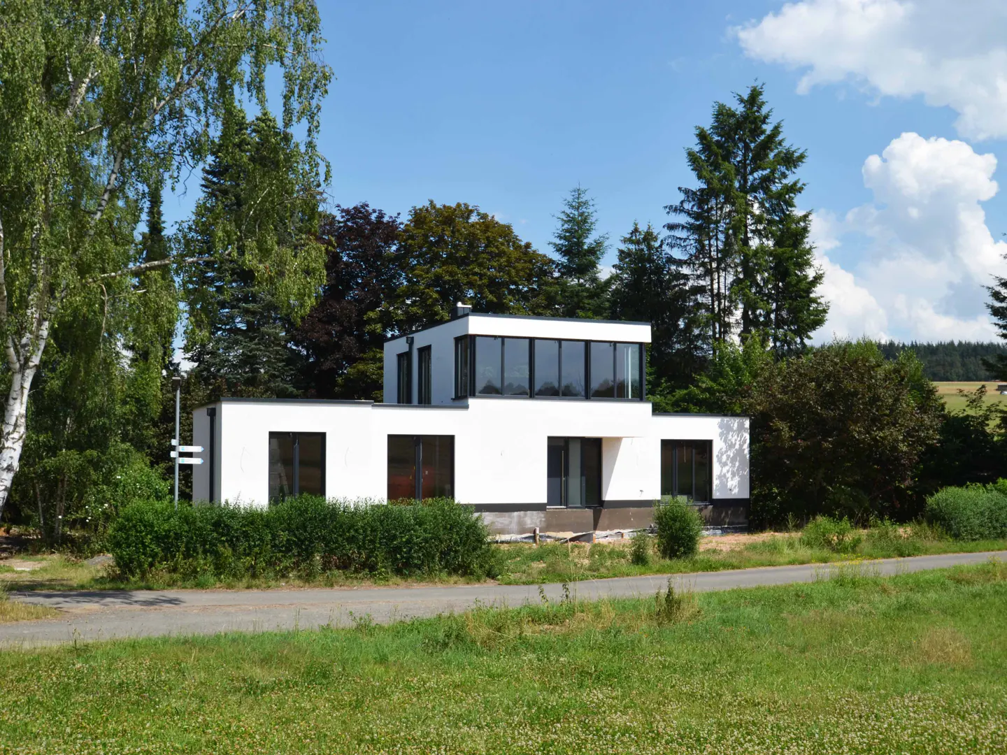 Modern white house with black framed windows, surrounded by green trees and grass under a blue sky.