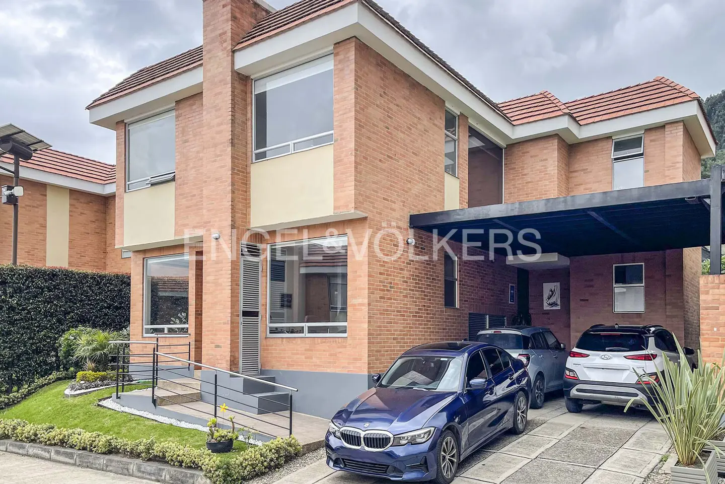 Two-story brick house with a red tile roof, a black carport, and three parked cars.