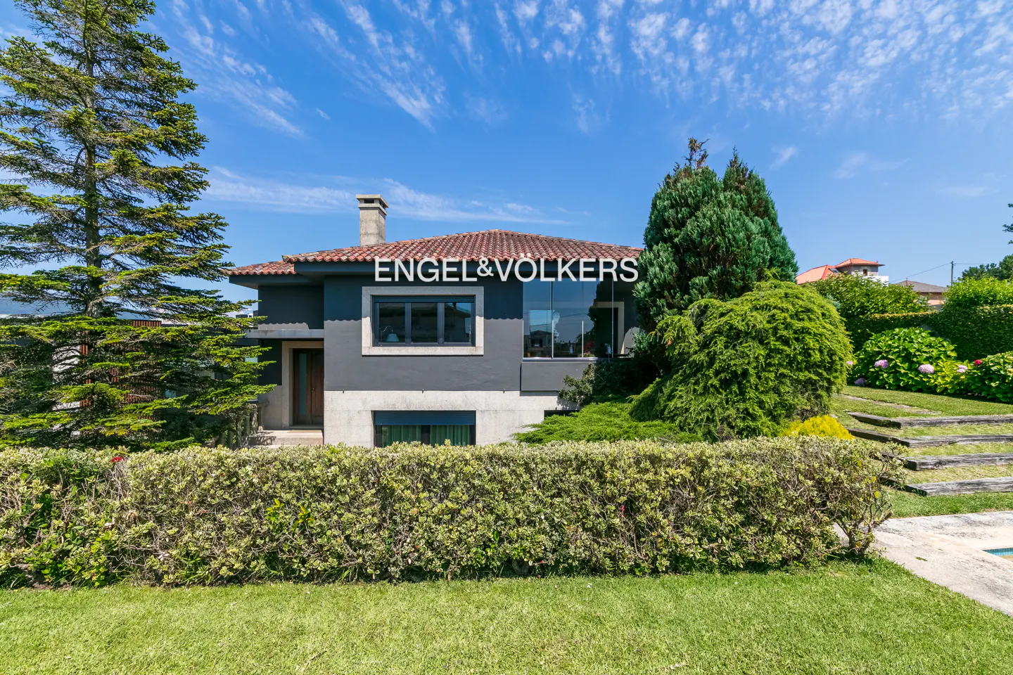Gray two-story house with a red tile roof, green lawn, and trimmed hedges under a blue sky. Engel & Volkers logo on the house.