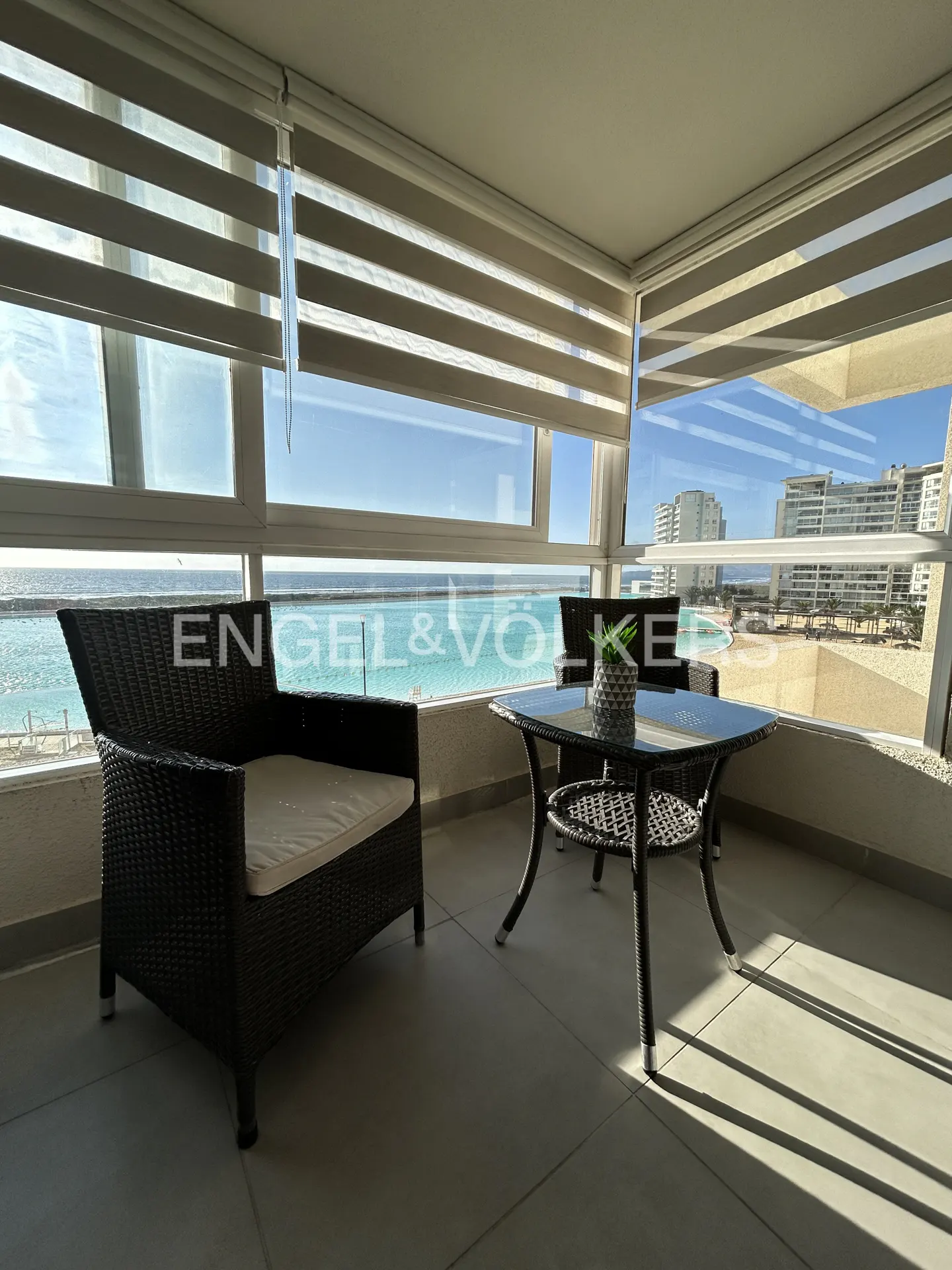 Sunlit balcony with black wicker chairs and a glass table overlooking a bright blue ocean and coastal buildings.