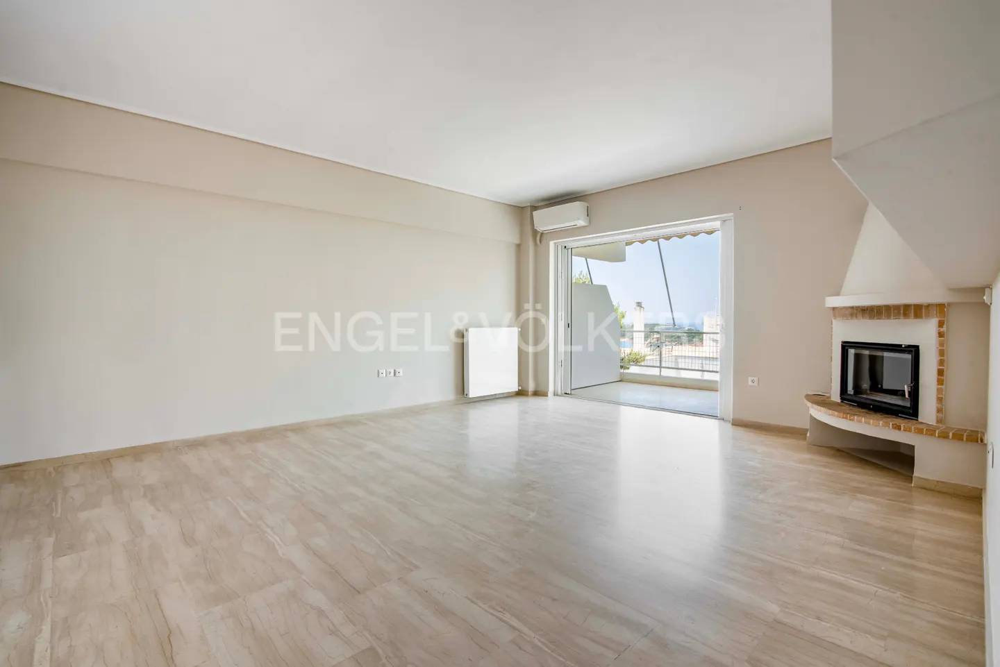 A bright, empty living room with beige walls, marble floors, and a fireplace. Balcony doors open to a view of the city.
