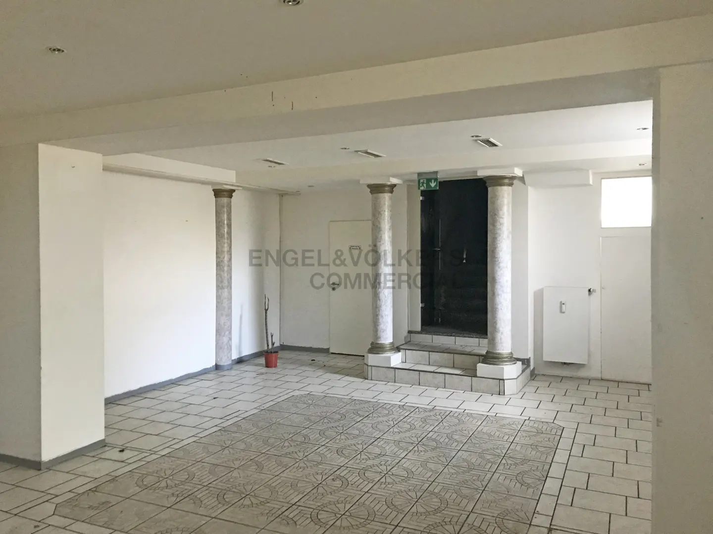 A bright, empty commercial space with white walls, tiled floors, and decorative columns. A black door with an exit sign is visible.