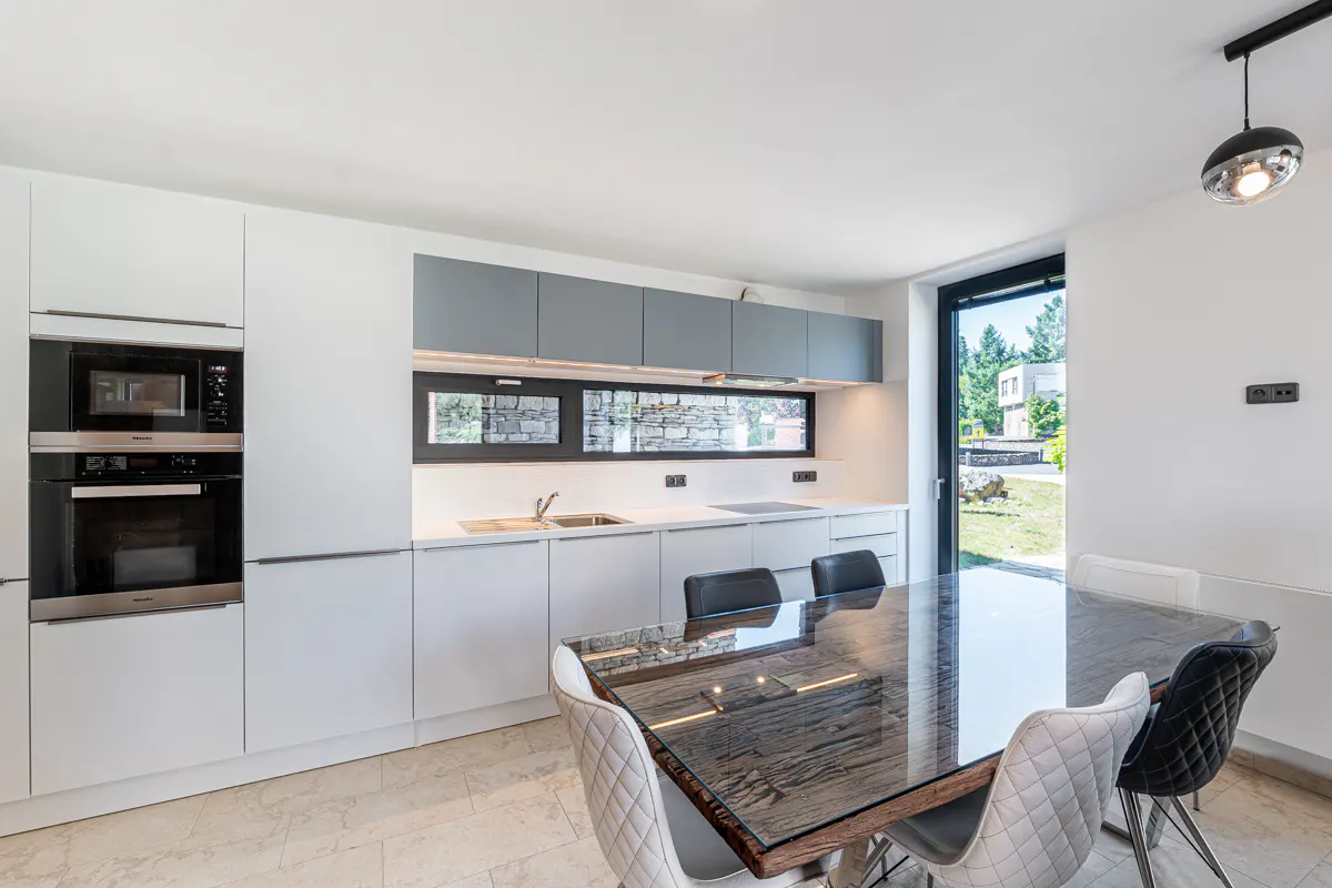 Bright kitchen with white cabinets, stainless steel appliances, and a dark wood table with upholstered chairs. A window overlooks a green yard.