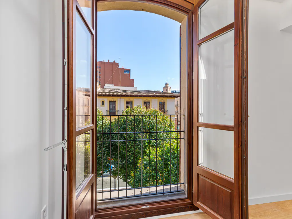 View from an open, dark wood framed window, showing a city street with trees and buildings under a blue sky.