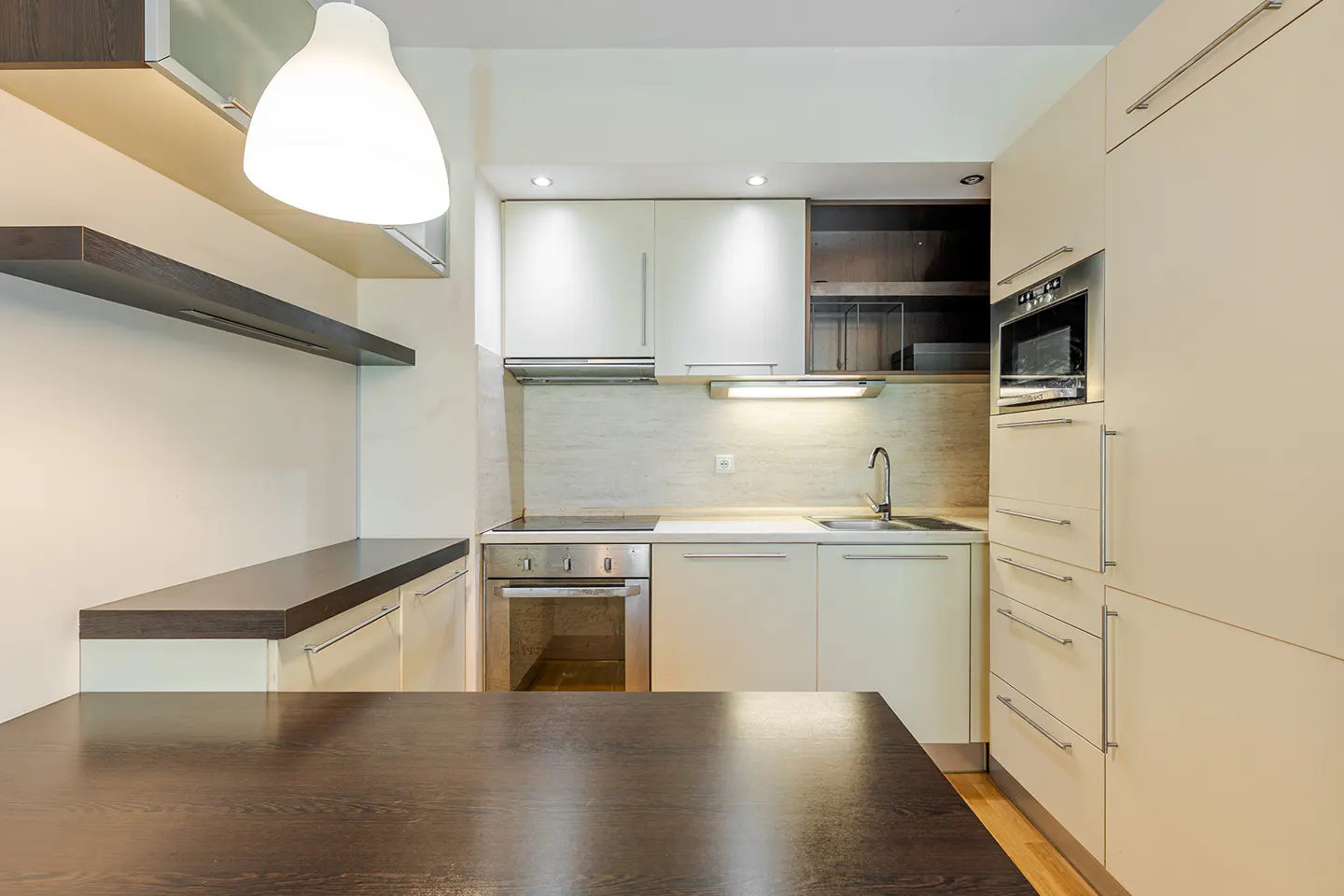 Modern kitchen with cream cabinets, stainless steel appliances, and dark wood countertops. A white pendant light hangs above the counter.
