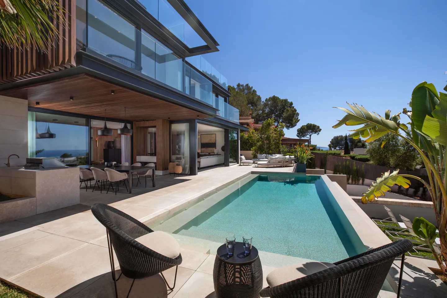Modern home exterior with a pool. Two black chairs face a small table with glasses. The house has wood accents and glass railings.