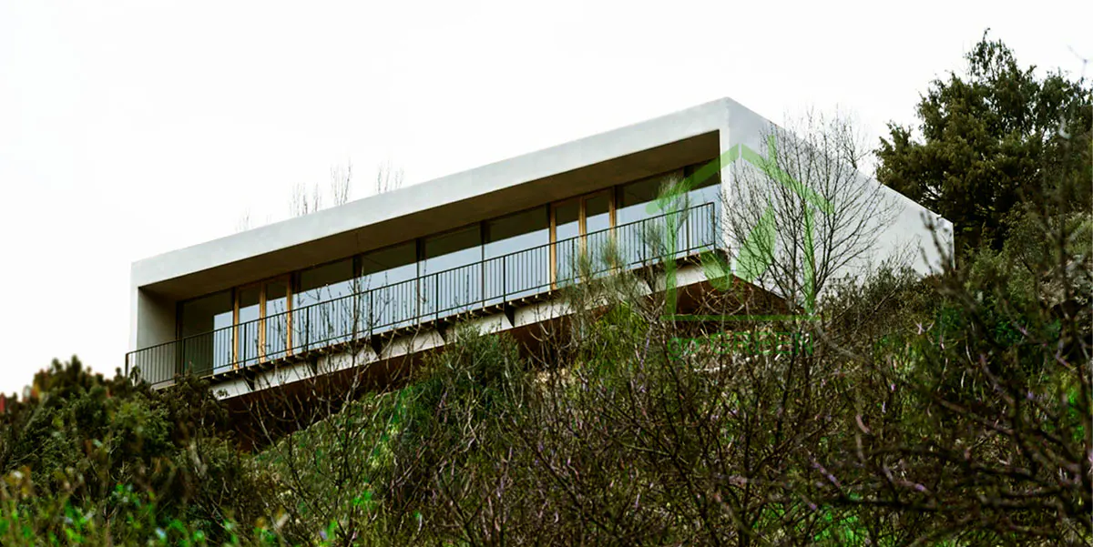 Modern white house with a balcony, set on a hill amongst trees and greenery.
